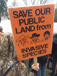 Person holding sign that reads "Save our public land from invasive species" with pictures of Bundy family